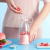 Woman preparing smoothie by adding strawberries to a pink portable electric juicer bottle